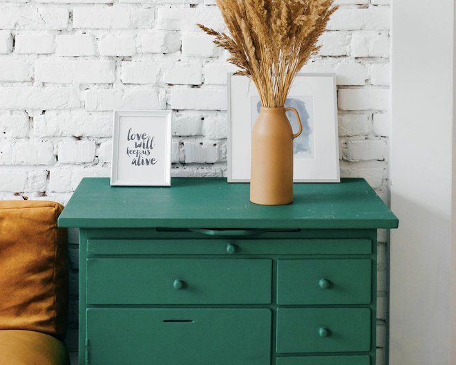 framed pictures and a tan vase with plants sit atop a rustic, wooden, green cabinet with drawers against a white brick wall